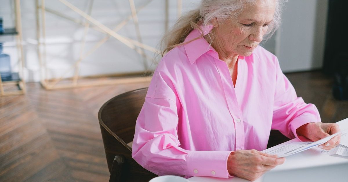 Mulher idosa de camisa rosa lendo papéis sobre uma mesa branca
